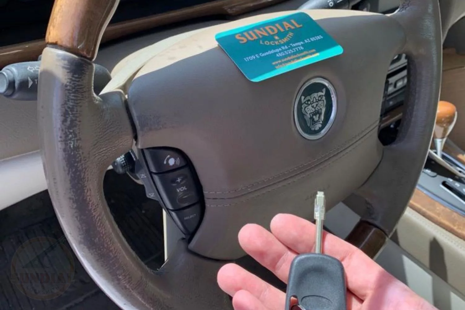 Close-up view of a Jaguar steering wheel with controls, with a 'SUNDIAL Locksmith' card placed on the dashboard and a hand holding a Jaguar car key in the foreground.