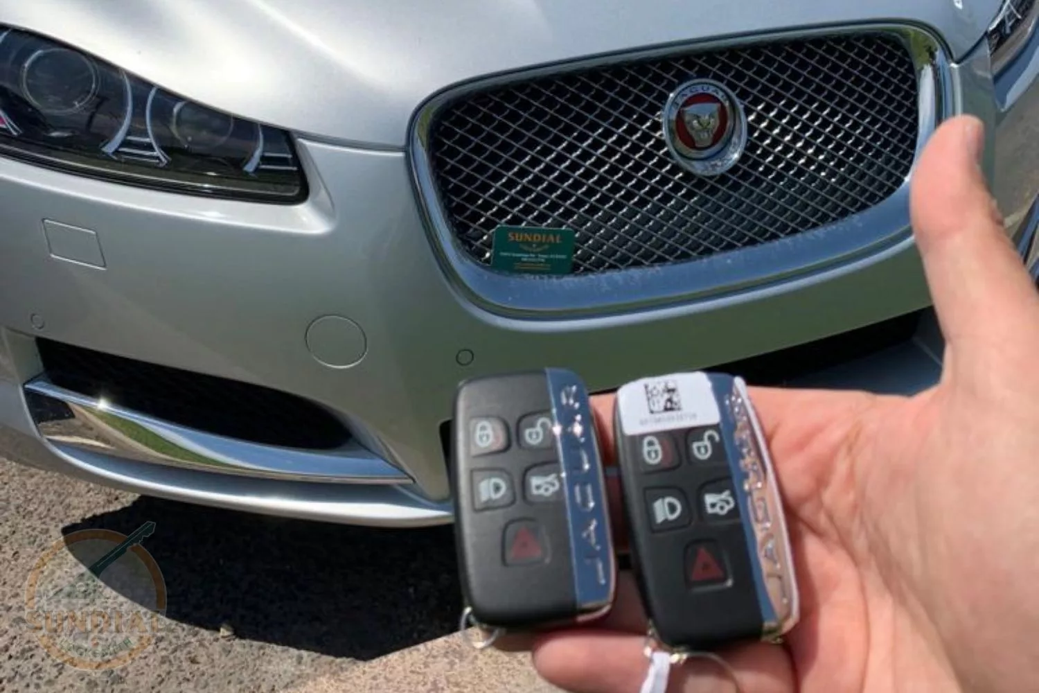 Hand holding two Jaguar key fobs in front of a silver Jaguar vehicle's front grille and headlight, with a 'SUNDIAL' business card placed on the grille