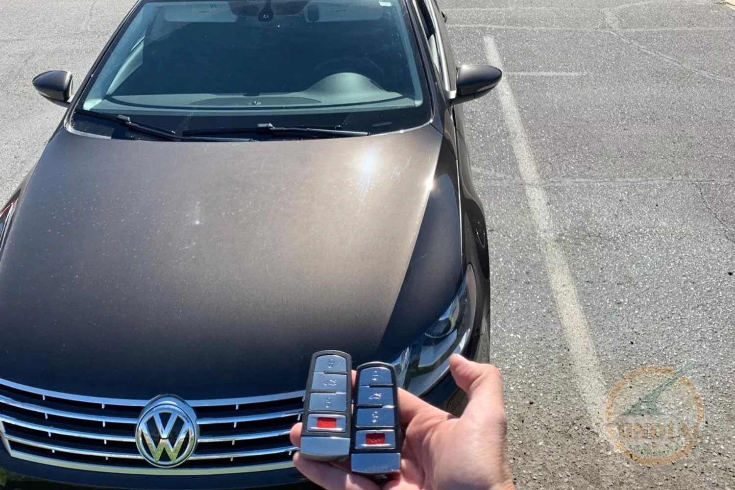 A person's hand holding two Volkswagen car keys in front of a parked Volkswagen car, with 'GROOMING' and 'LOCKSMITH' store signs in the background.