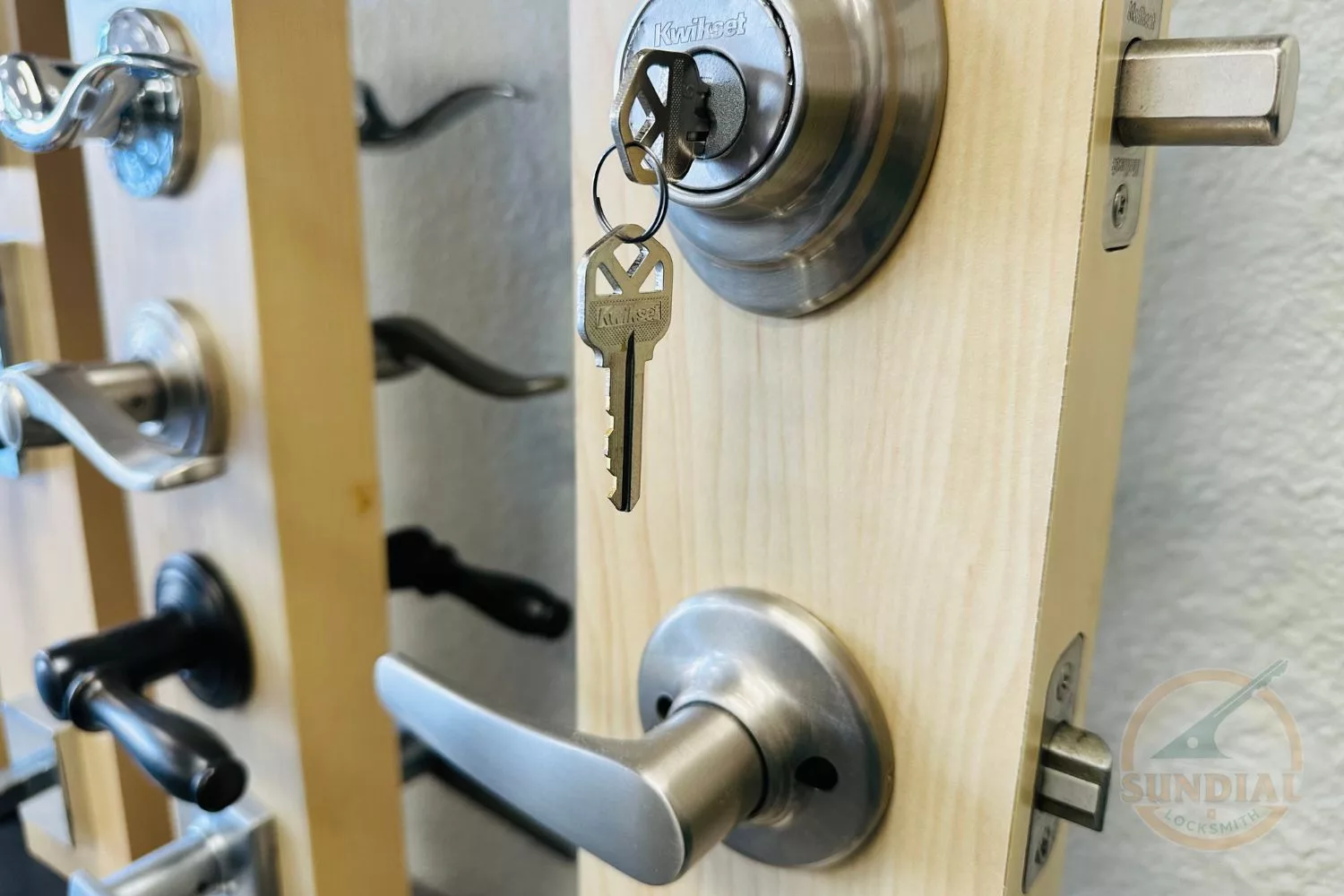 Close-up view of a door handle and keyhole with a 'Kwikset' key inserted, surrounded by various door handles and hardware, with the 'Sundial Locksmith' logo watermark.