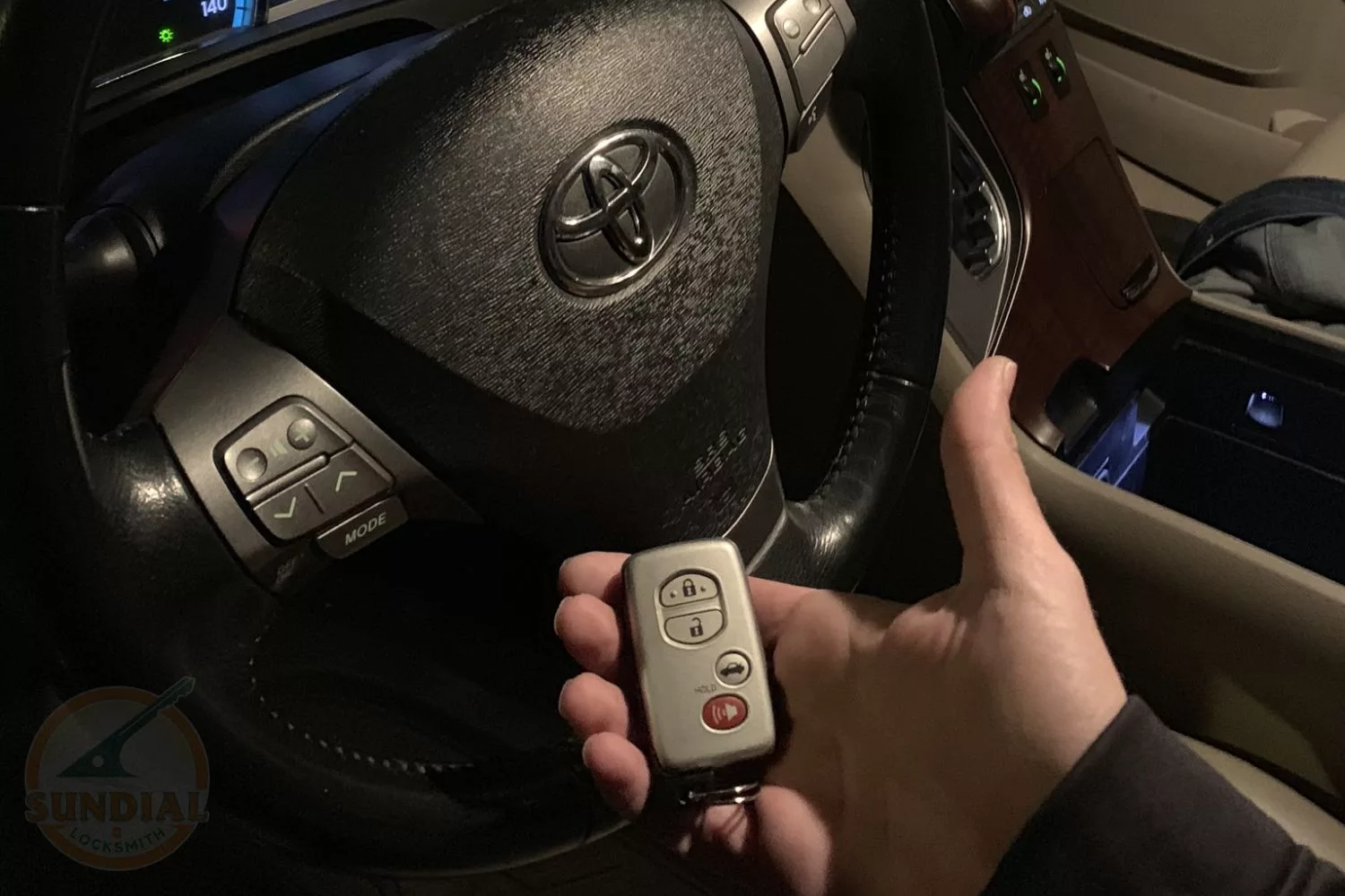 Hand holding a car key fob in front of a Toyota vehicle's steering wheel and dashboard at night.