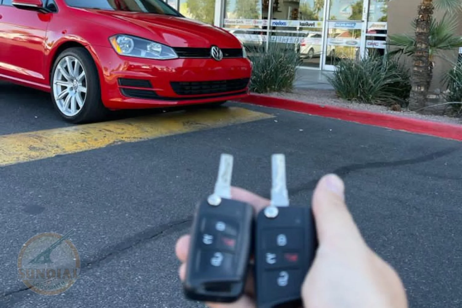 A hand holding two Volkswagen key fobs in front of a red Volkswagen Golf parked outside a locksmith shop.