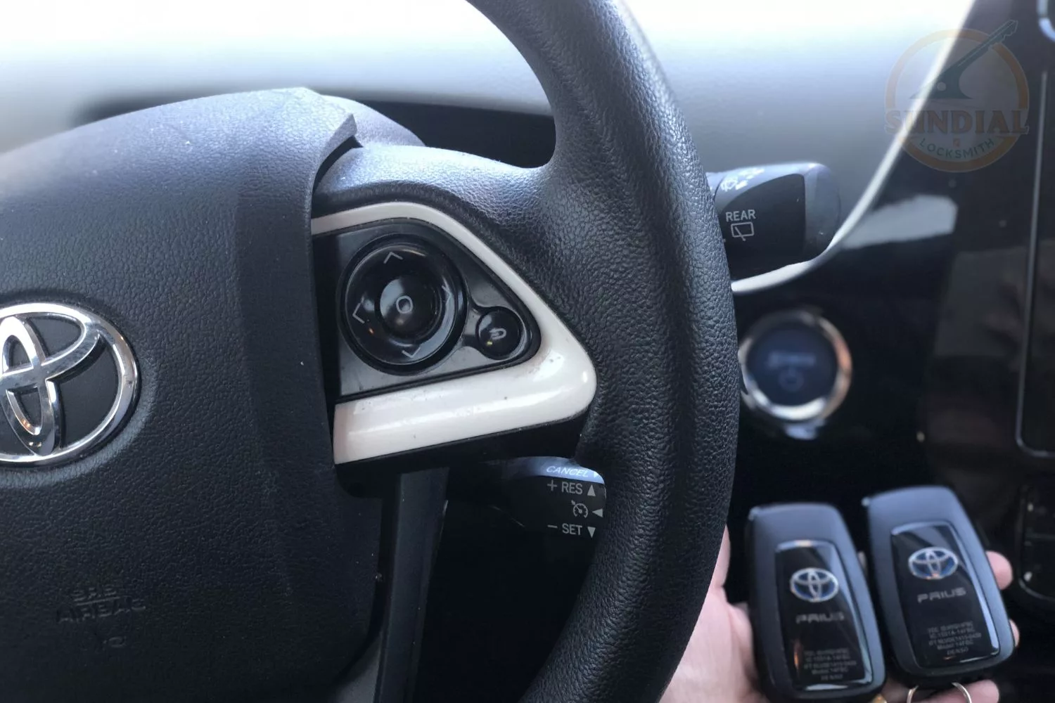 Close-up of a Toyota steering wheel with cruise control buttons and two Toyota Prius key fobs held in front.