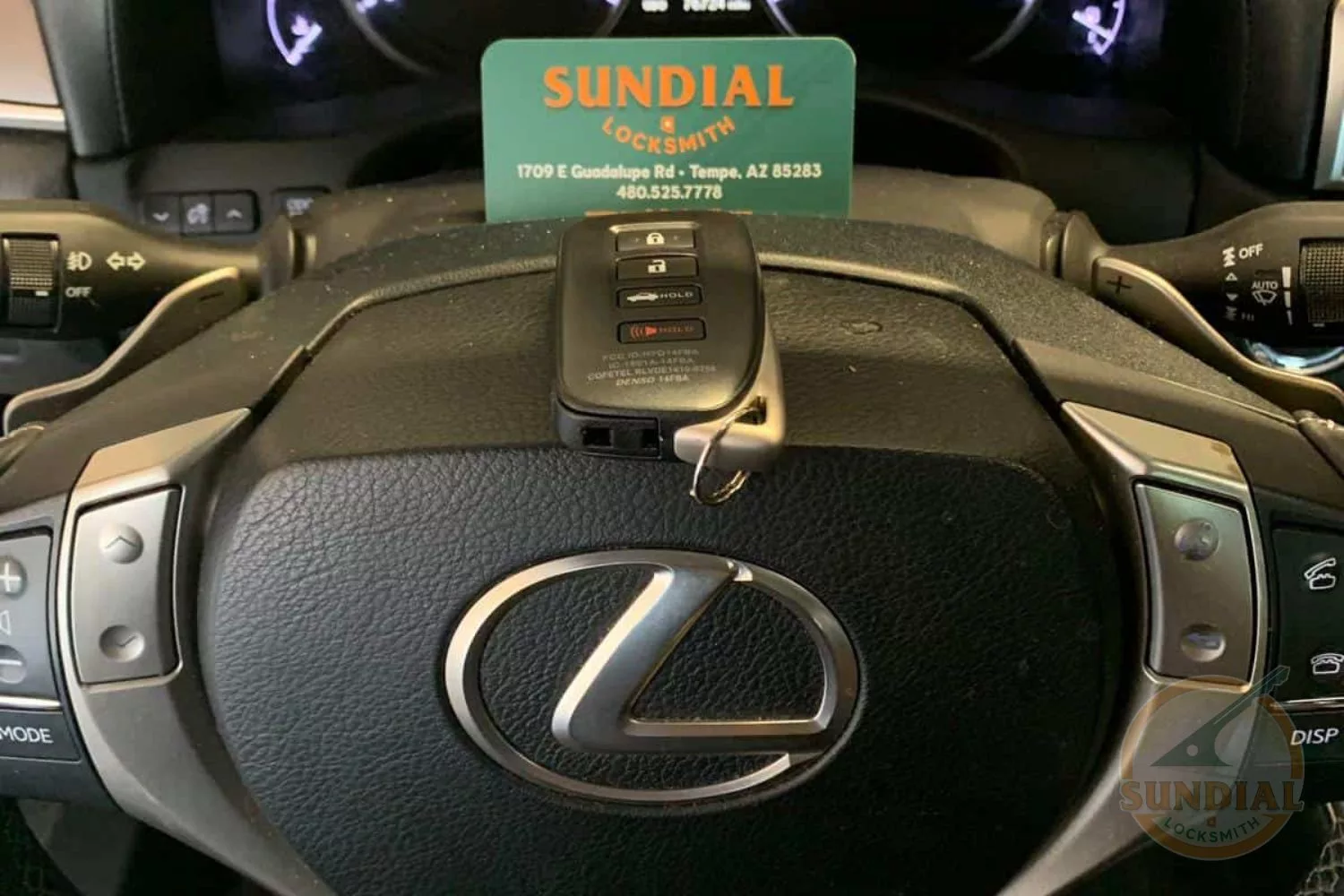 A Lexus car key placed on the steering wheel, with a Sundial Locksmith business card propped behind it.
