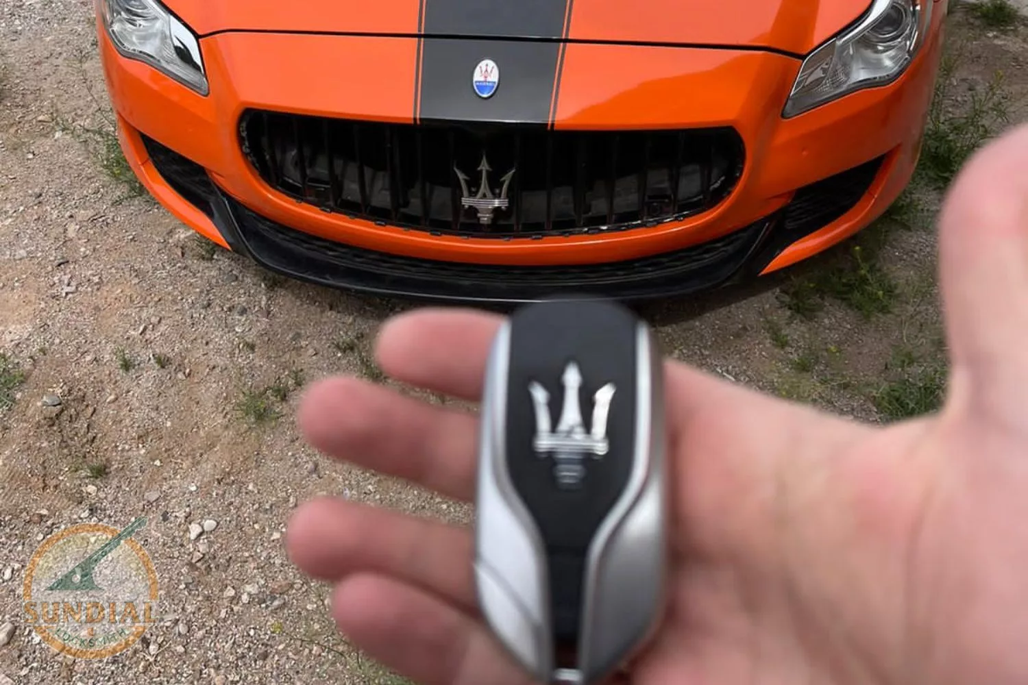 A hand holding a Maserati car key with an orange Maserati vehicle in the background.