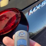 Close-up of a hand holding a new Mazda key fob with the MX-5 model emblem in the background.
