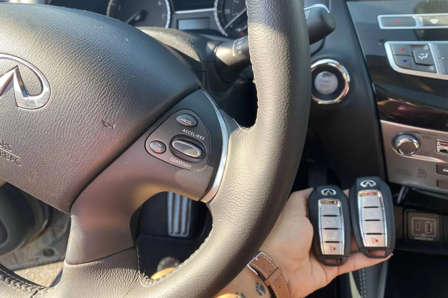A person's hand holding two key fobs with a luxury vehicle's steering wheel and dashboard controls in the background.