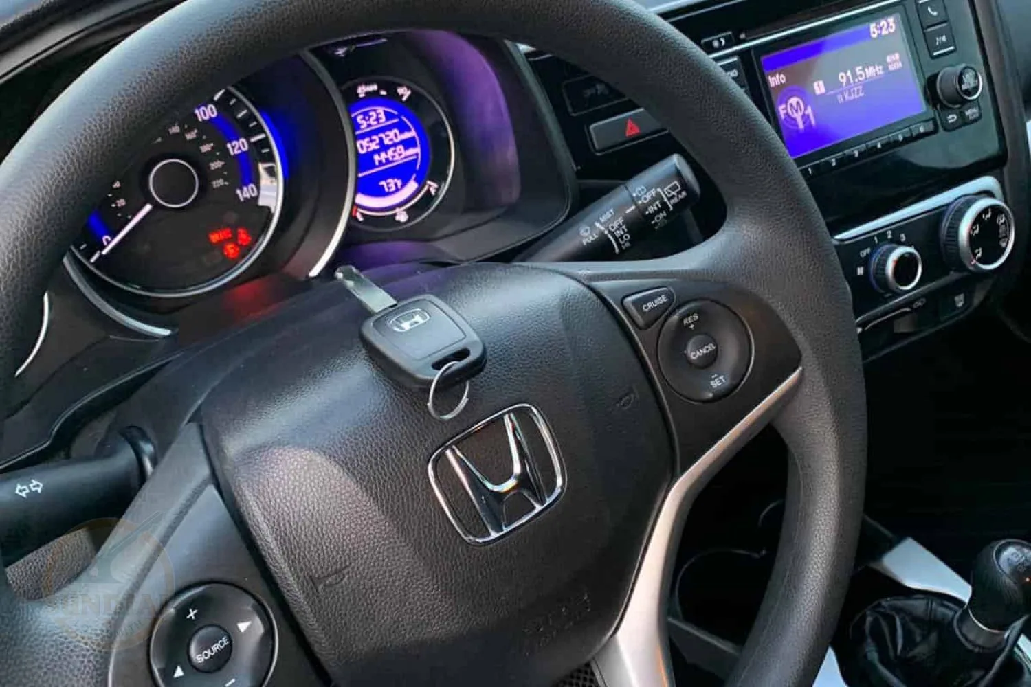 A Honda car key inserted into the ignition of a vehicle, with the steering wheel, gauge cluster, and multimedia system in the background.