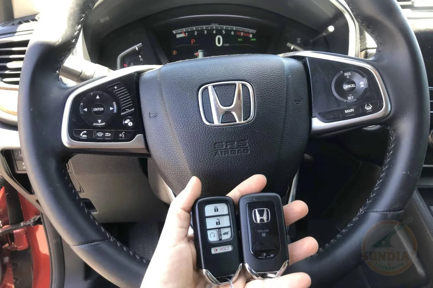 A person's hand holding two Honda car key fobs in front of the steering wheel with mounted audio and cruise control buttons.