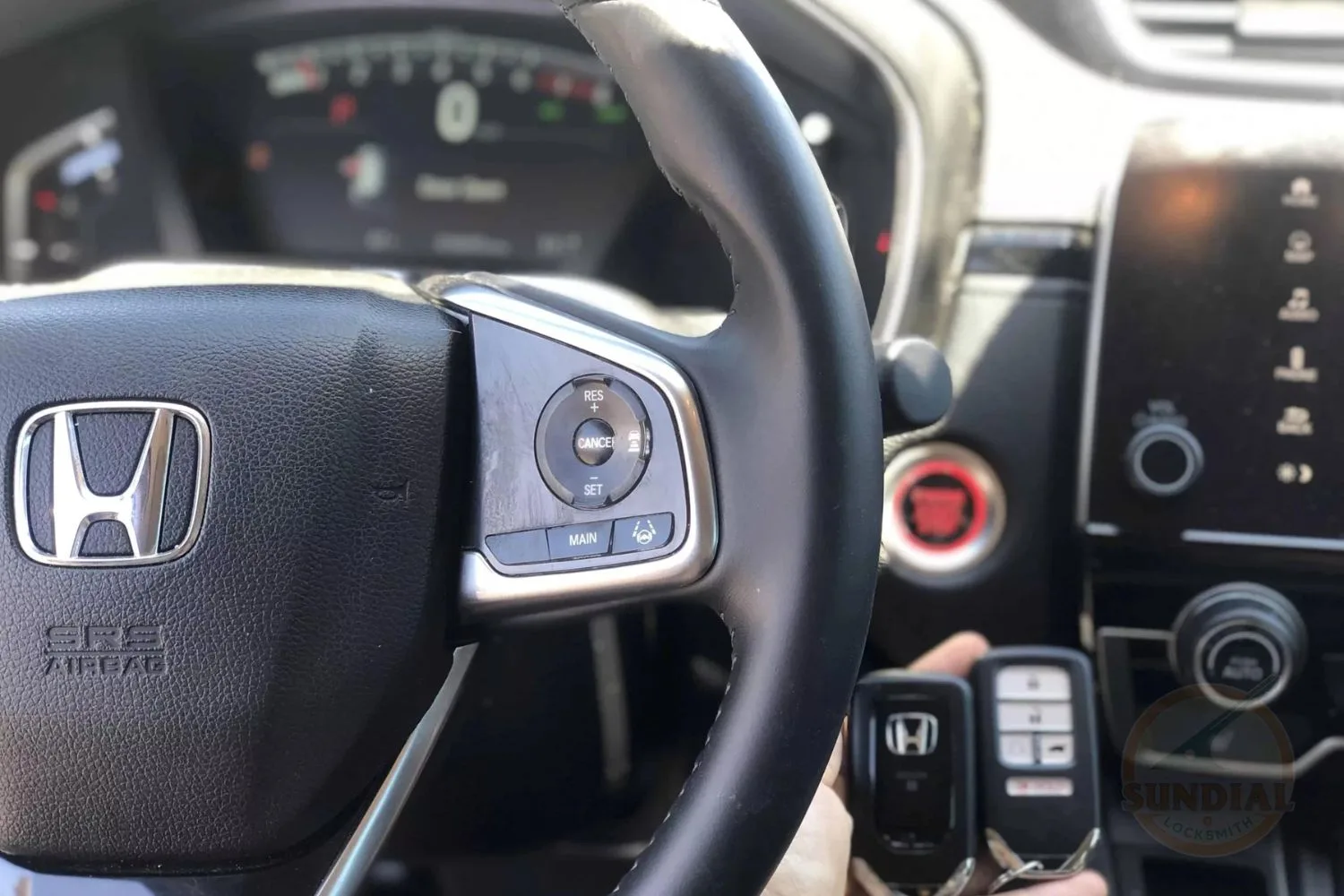 A Honda key fob held in the foreground with the steering wheel and vehicle control buttons of a Honda car in the background.