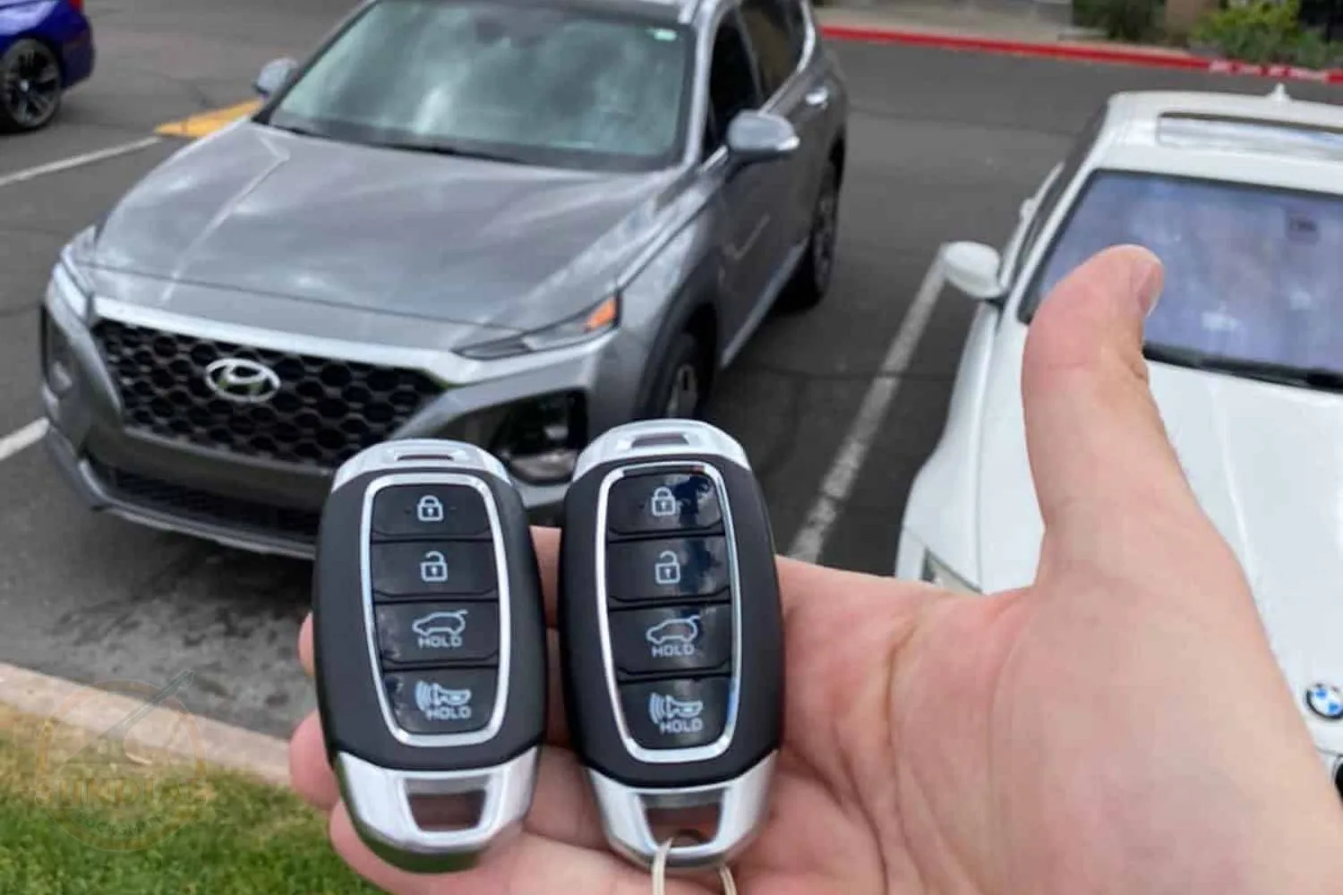 A person's hand holding two Hyundai car key fobs in a parking lot, with a grey Hyundai SUV in the background.
