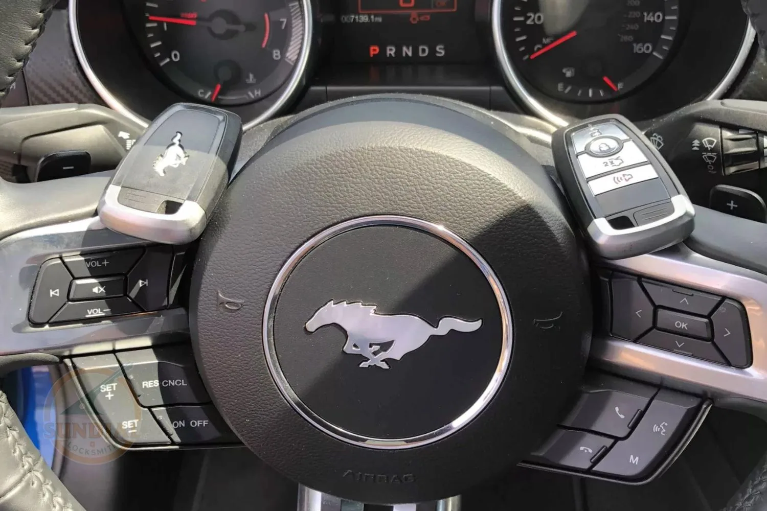 A close-up view of two Ford Mustang key fobs on the steering wheel with audio and cruise control buttons, against the backdrop of the car's instrument cluster.