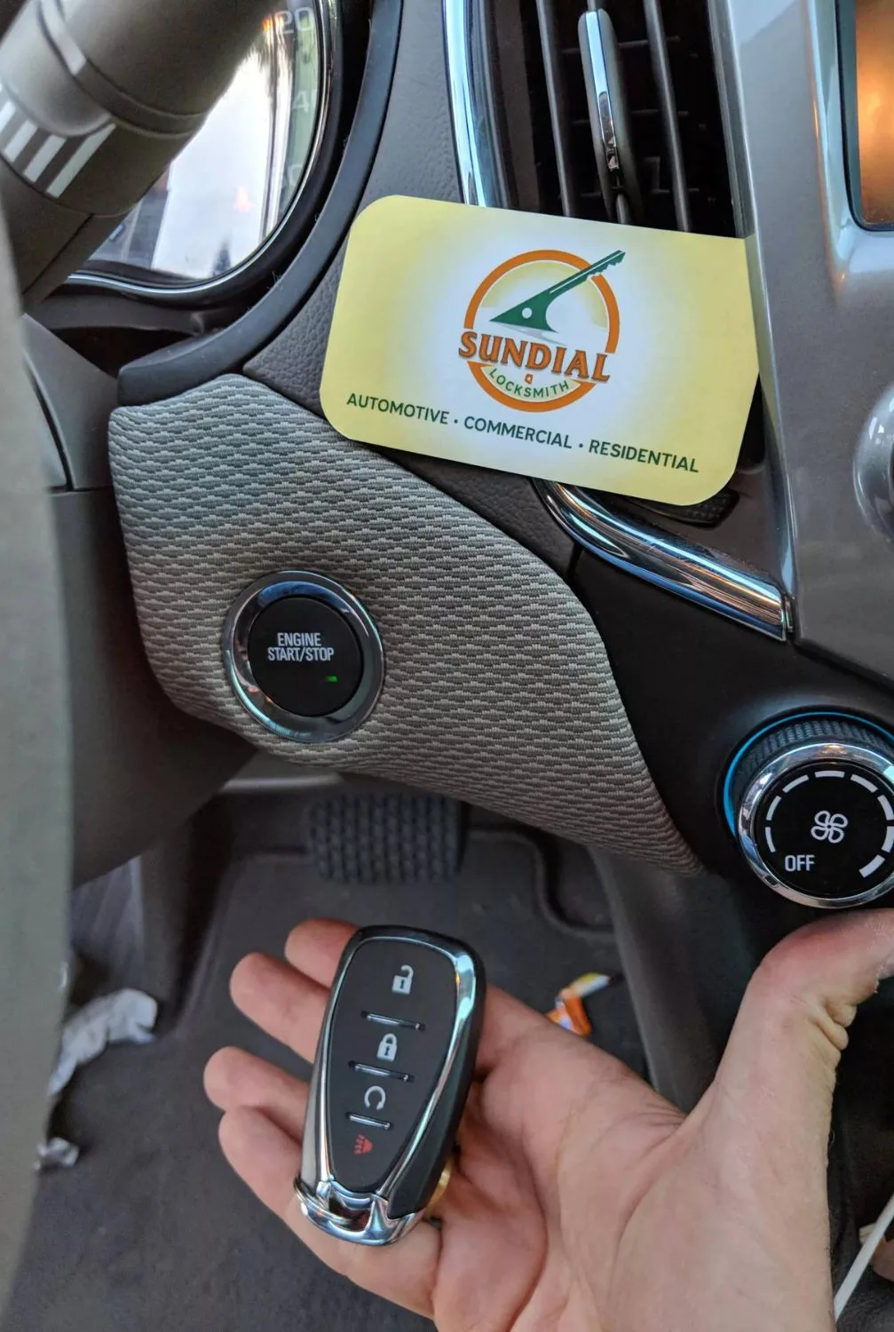 A hand holding a car key fob with a push-to-start button and a business card on the dashboard.