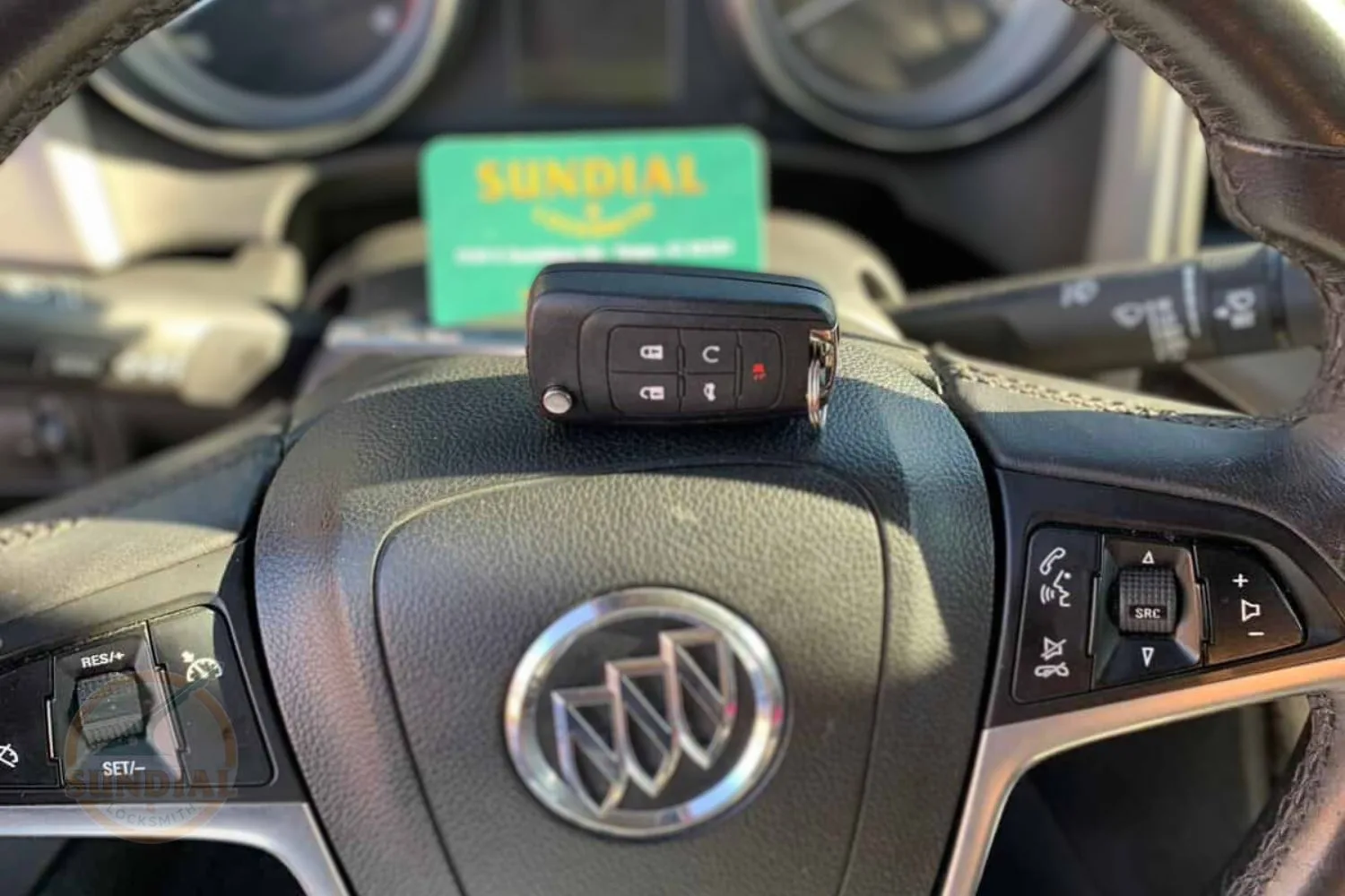 Buick car key fob held in front of the steering wheel with the Sundial Locksmith business card displayed behind it.