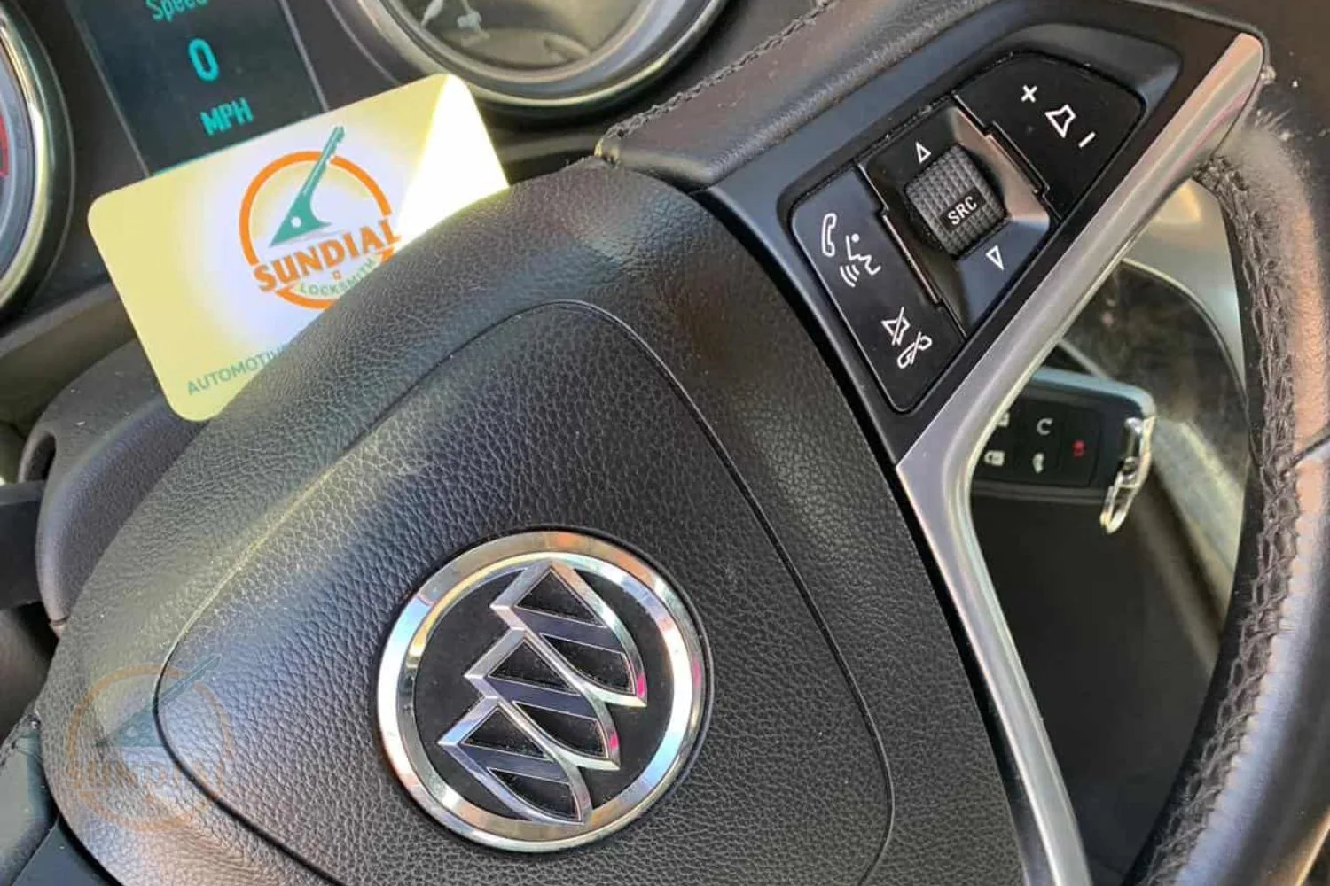 Buick car key fob placed over the steering wheel emblem with a Sundial Locksmith business card in the dashboard slot.