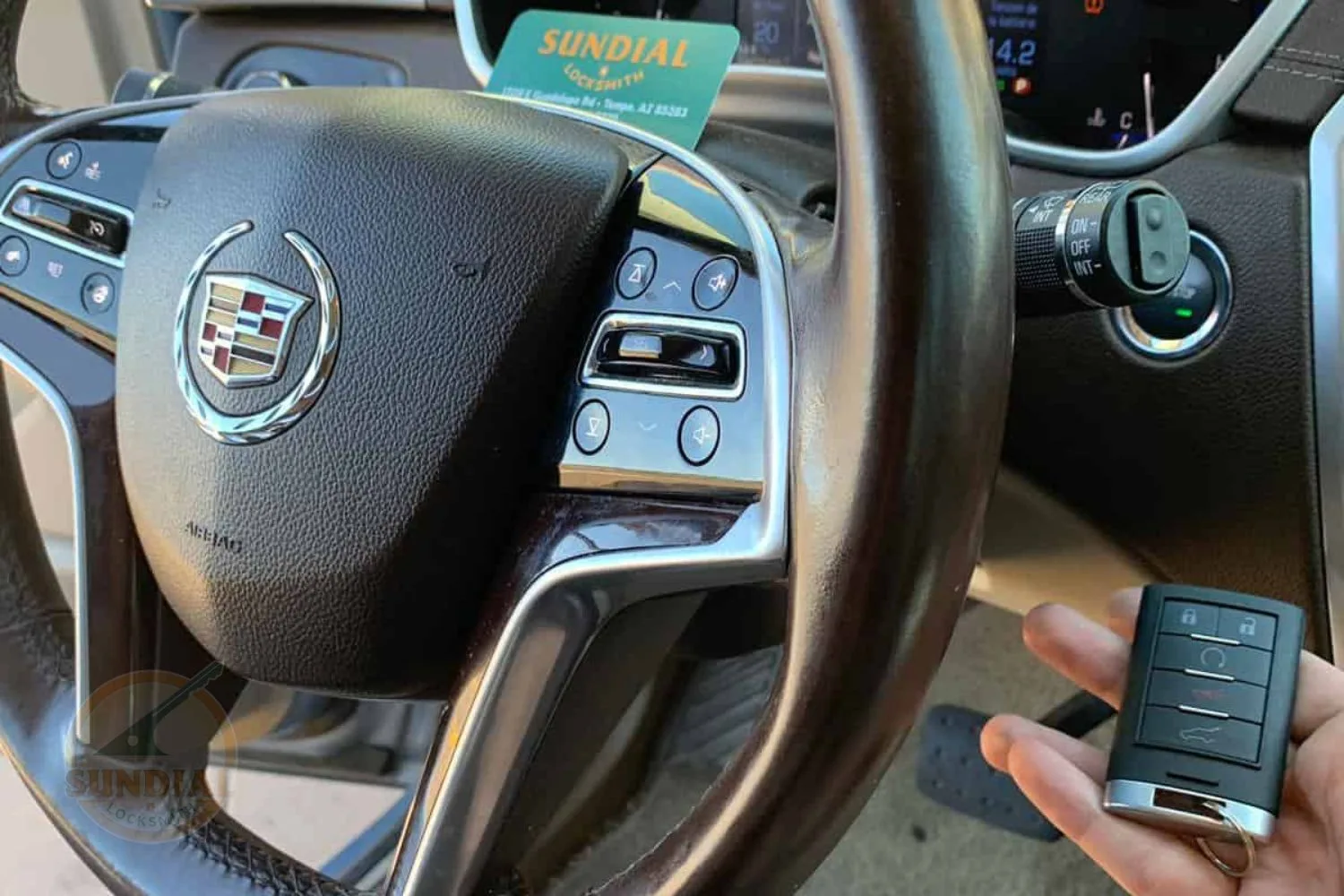 Cadillac car key fob in hand with the steering wheel and dashboard in the background, featuring the Sundial Locksmith business card.