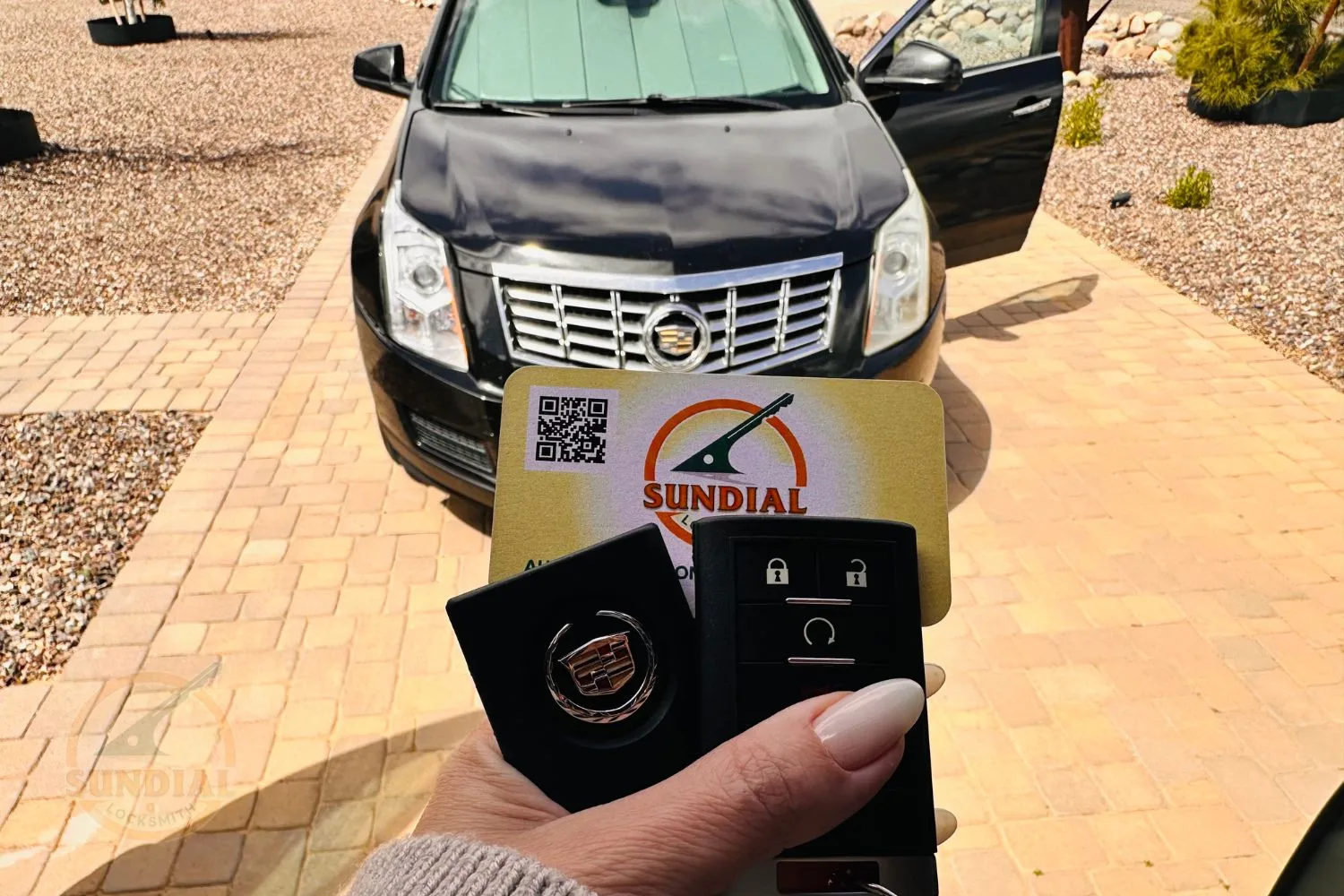 A person's hand holding a Cadillac key fob in front of a Cadillac car, with a Sundial Locksmith business card displayed prominently.