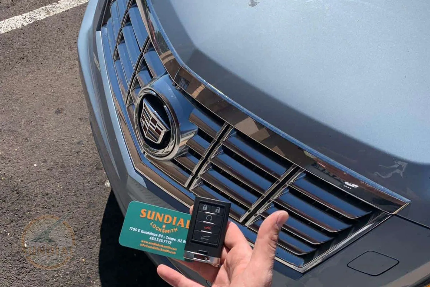 A Cadillac car key fob held in a hand in front of the vehicle's emblem and grille, with a Sundial Locksmith business card visible