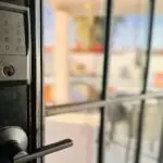 A close-up of a black keypad door lock installed on a glass pane door, with a blurred indoor background.