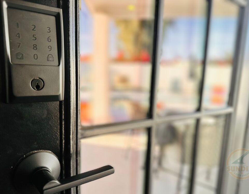 A close-up of a black keypad door lock installed on a glass pane door, with a blurred indoor background.