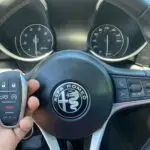 A close-up of an Alfa Romeo steering wheel with a hand holding the car's key fob in front.