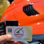 A hand holding a Sundial Locksmith business card and two car key fobs in front of a bright orange car.