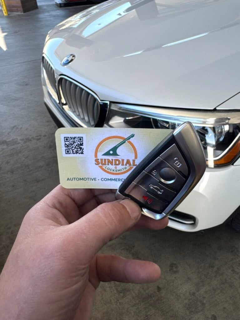 A hand holding a BMW smart key fob and a Sundial Automotive Locksmith business card in front of a white BMW vehicle.