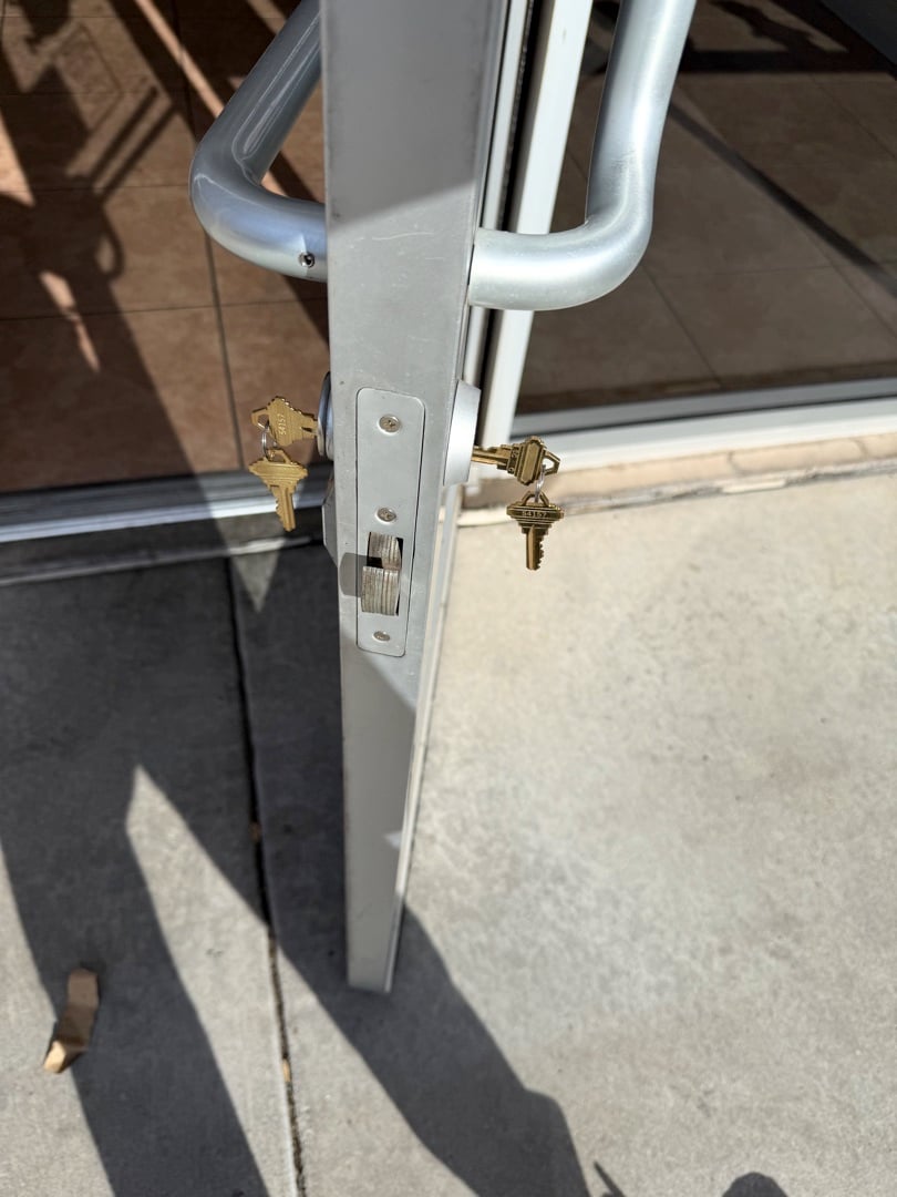 A close-up view of a sliding screen door's latch mechanism with two keys inserted into separate locks on an aluminum door frame.