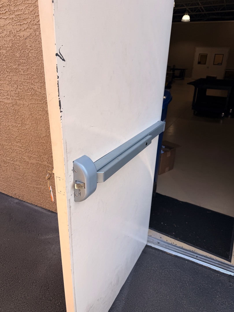 Panic Bar Installation: What Every Business Owner Needs to Know A silver push bar panic hardware exit device installed on a white commercial metal door, viewed from the interior side of a building.