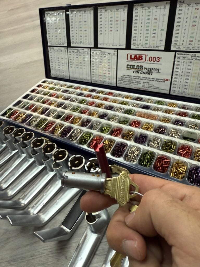 Can a Locksmith Rekey a Car's Ignition? Here's the Honest Answer A locksmith holds a brass key and lock cylinder in front of an open LAB .003 pin kit tray filled with colorful assorted lock pins organized in rows.