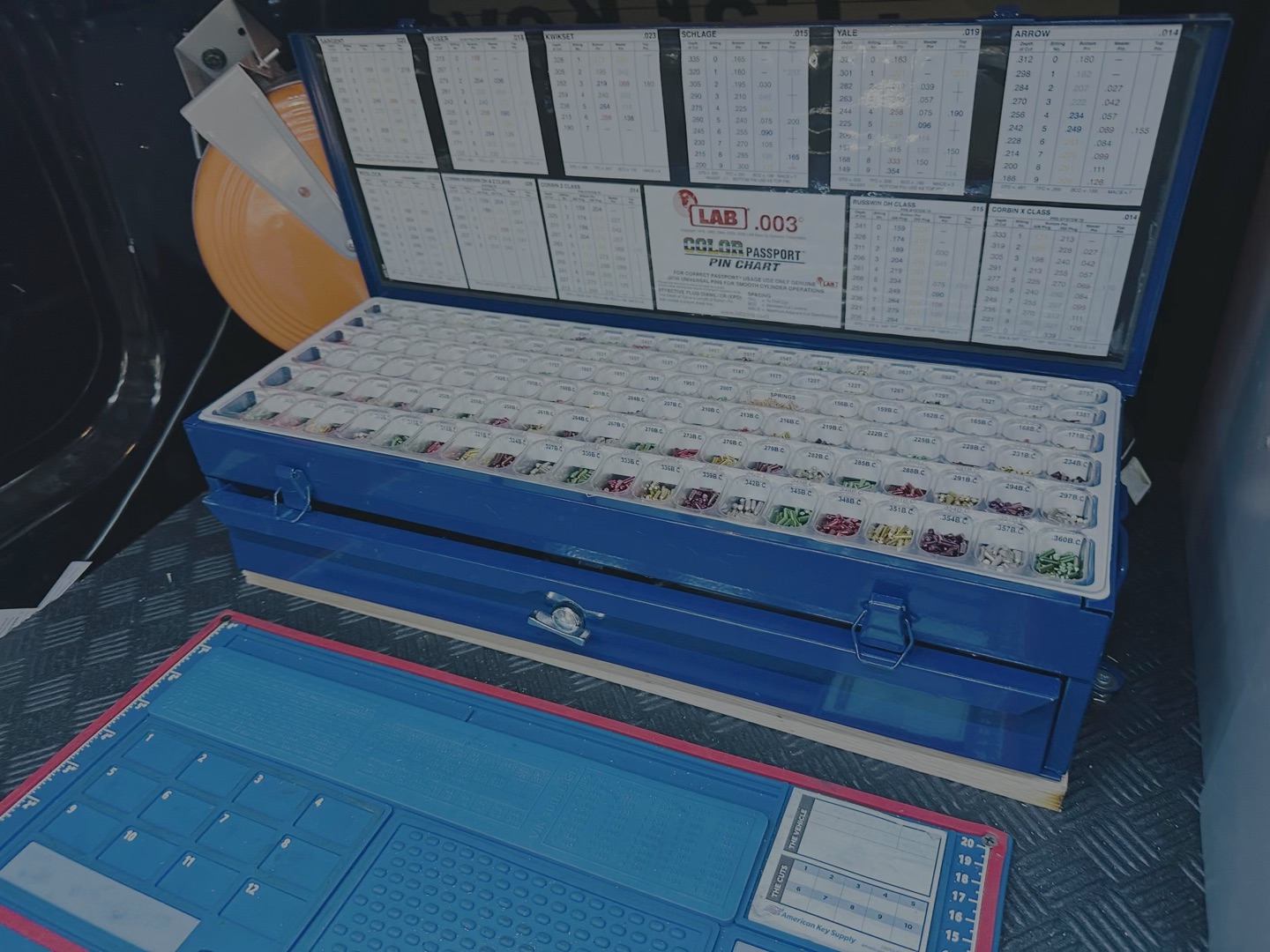 A blue locksmith case open in a vehicle trunk, filled with rows of key blanks and code reference charts, alongside a blue laptop.