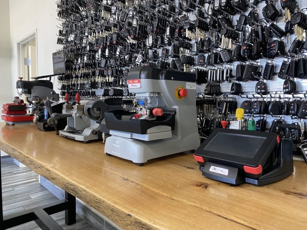 How Many Key Fobs Can Actually Be Programmed to One Car? Three professional key cutting machines lined up on a wooden workbench inside a locksmith shop, with a large wall display of key blanks and fobs in the background.