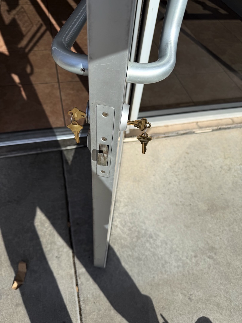 Close-up of a sliding glass door edge showing two brass keyed lock cylinders installed in a silver aluminum door frame.