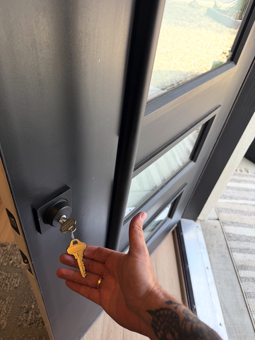 A tattooed hand holds a yellow key near the lock of a dark gray front door with a glass panel.