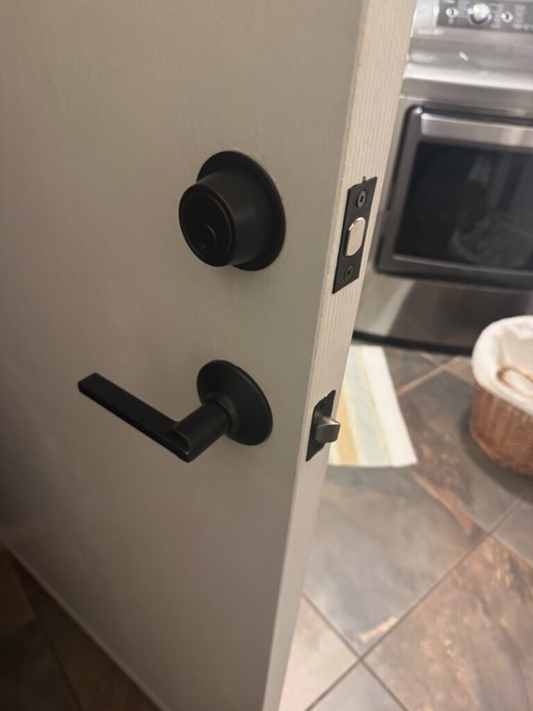 A residential interior door featuring a newly installed matte black deadbolt and lever-style door handle set on a white door frame with tile flooring visible in the background.