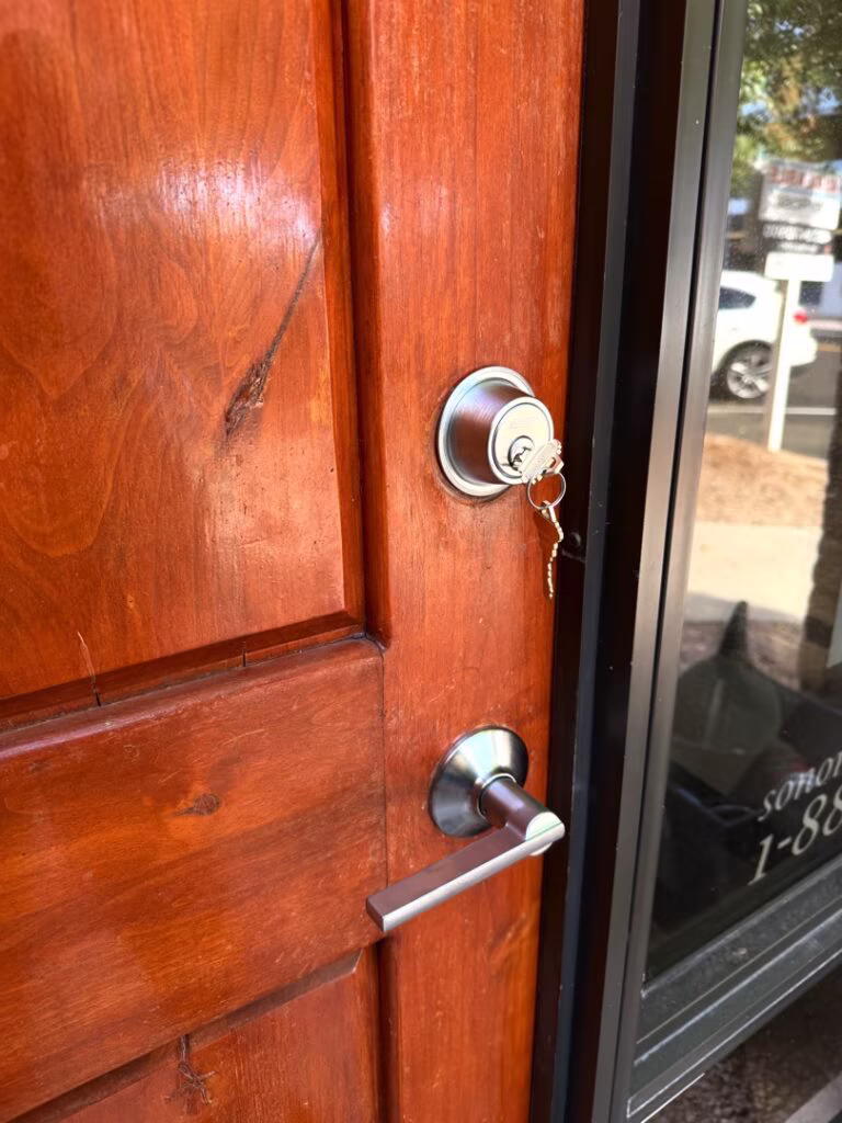 A silver deadbolt lock with keys inserted sits above a lever handle on a solid wood entry door with a glass sidelight panel.