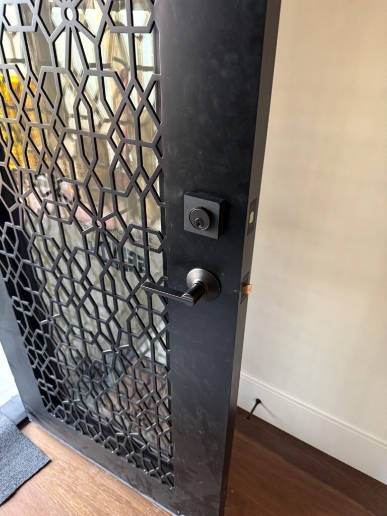 Grade 1 Deadbolt Installation: What It Is and Who Actually Needs One A dark metal security door featuring an ornate geometric lattice pattern, fitted with a satin nickel deadbolt and door knob hardware on a residential entryway.