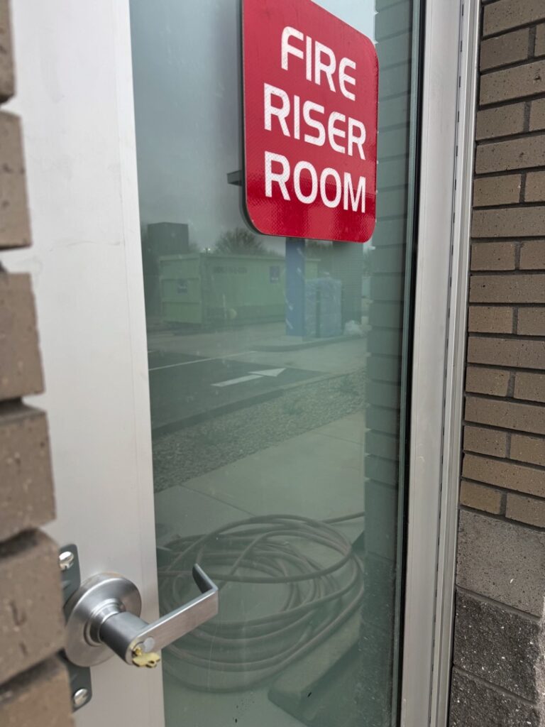 Access Control for an Office Suite: What to Expect from Installation A glass-panel commercial door with a red Fire Riser Room sign and a silver lever handle, located at the entrance to a building utility room.