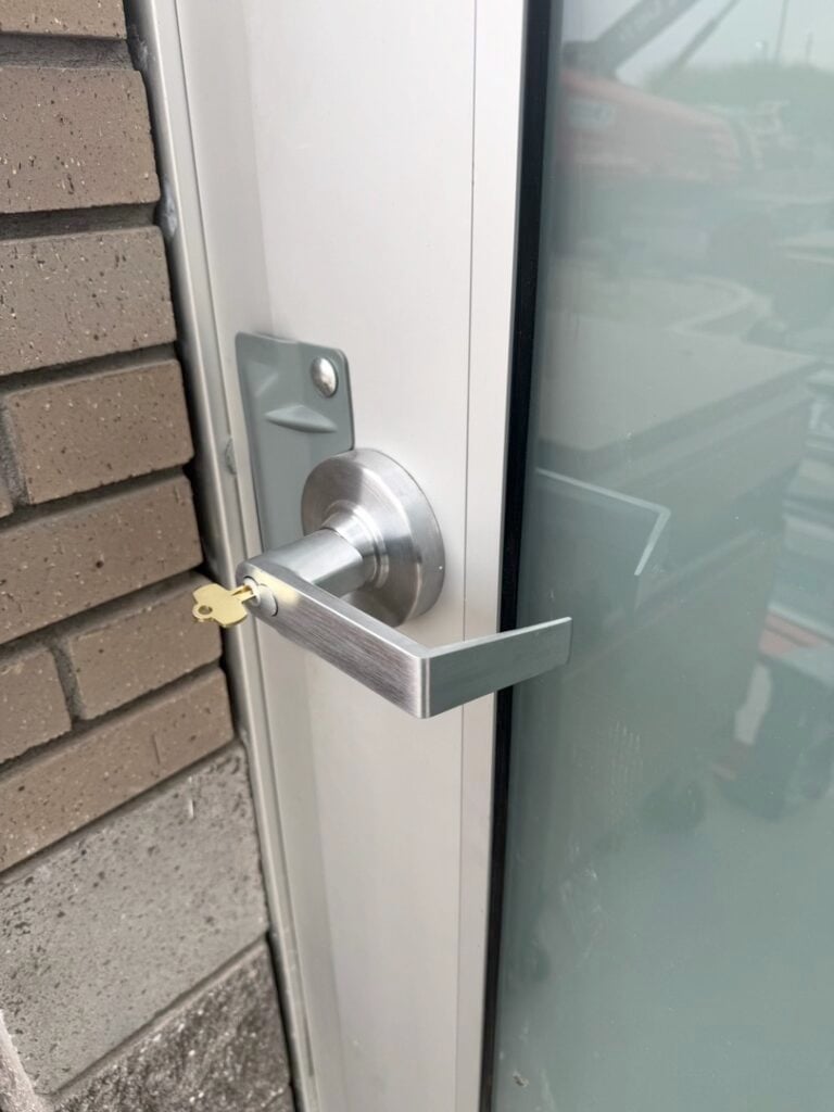 Commercial Door Lever Handle Lock Installation A brushed silver lever door handle and lock cylinder installed on a white aluminum commercial door frame next to a brick wall and glass panel.
