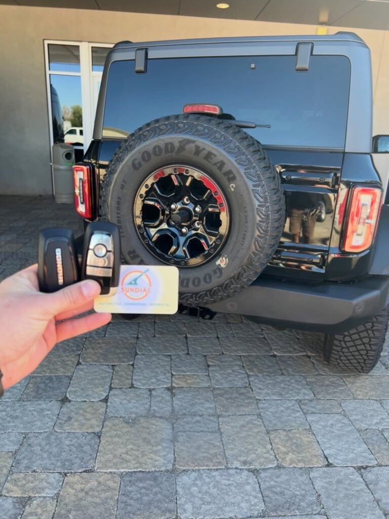A hand holding a new key fob and smart key next to the rear spare tire of a dark blue Jeep Wrangler in a paved driveway.