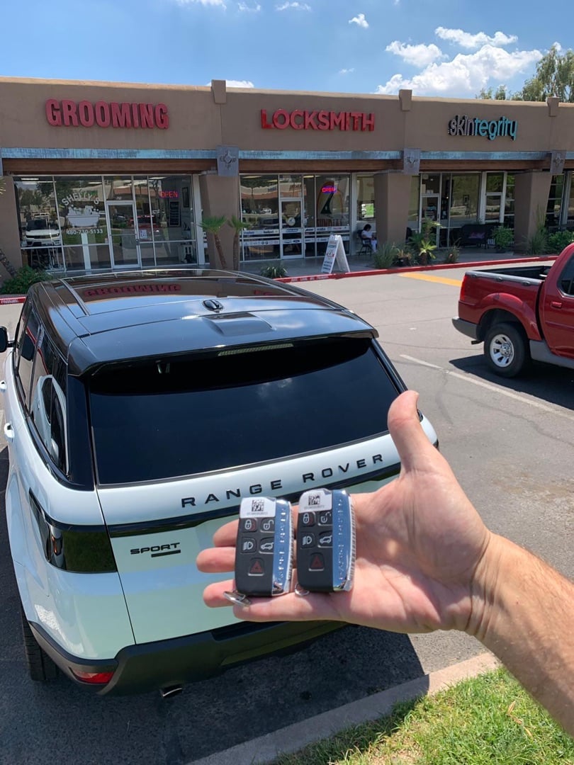 A hand holding two Range Rover key fobs in a parking lot in front of a strip mall with a white Range Rover Evoque parked nearby.