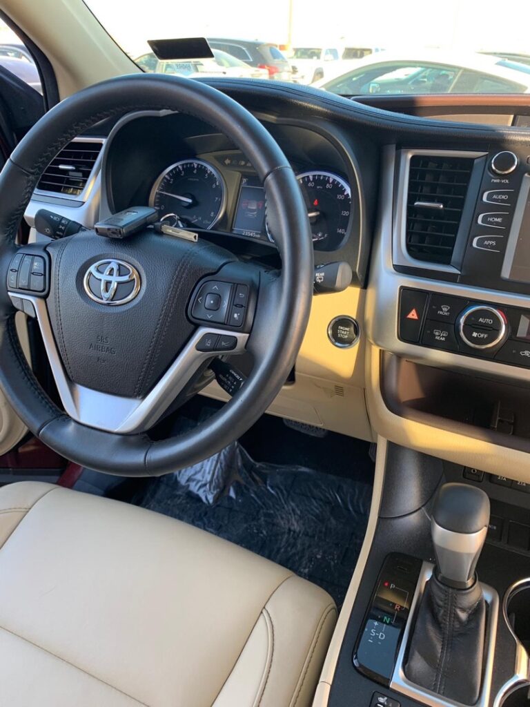 Interior view of a Toyota SUV showing the steering wheel, instrument cluster, center console with automatic gear shifter, and beige leather seats.