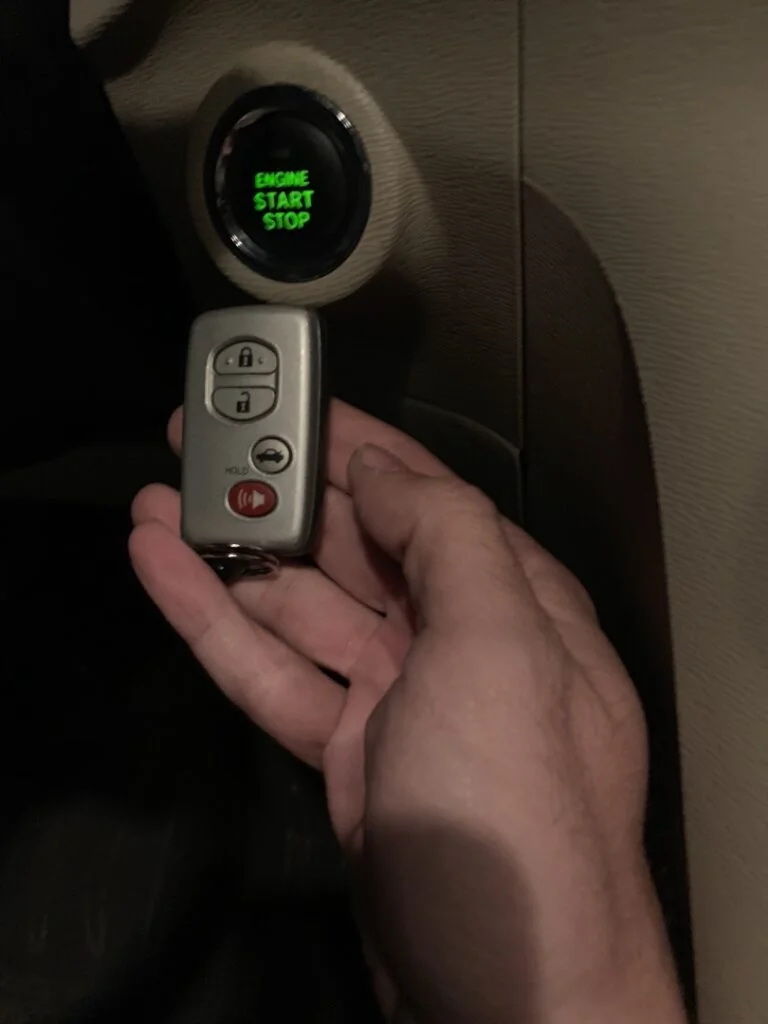 Signs Your Ignition Cylinder Is Failing Before It Strands You A hand holding a Toyota smart key fob next to a glowing green engine start stop push button ignition inside a vehicle.