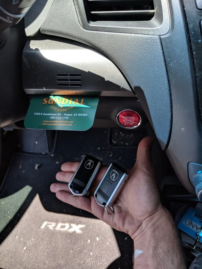 A locksmith holds a new Acura key fob next to the push-button ignition of an Acura RDX dashboard, with a Sundial Locksmith business card visible in the background.