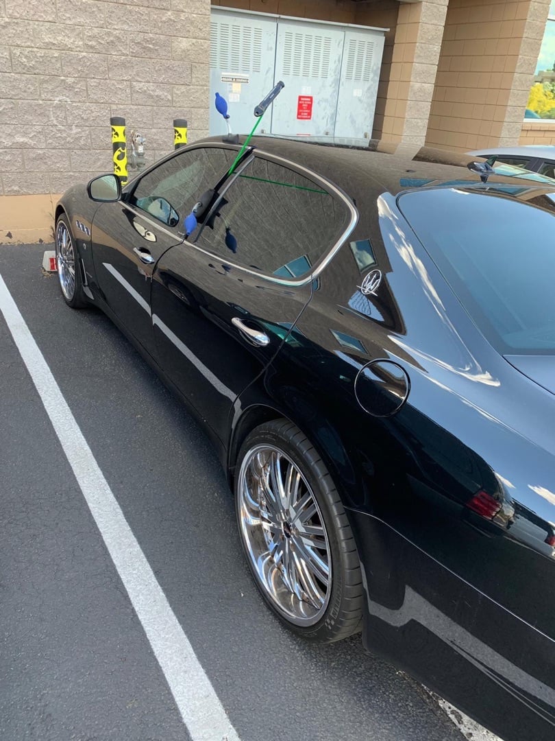 A shiny black luxury sedan parked in a parking lot with yellow locksmith slim jim tools visible near the door and window.
