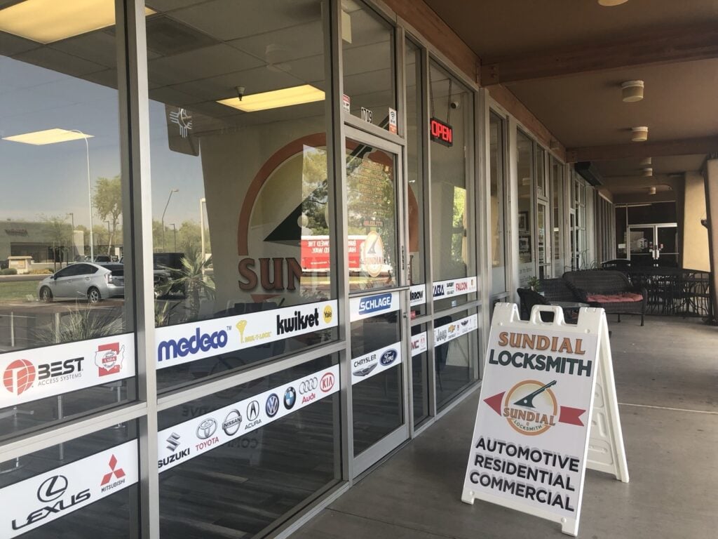 Exterior view of Sundial Locksmith shop front featuring glass windows covered with automotive brand decals and a sandwich board sign advertising automotive, residential, and commercial locksmith services.