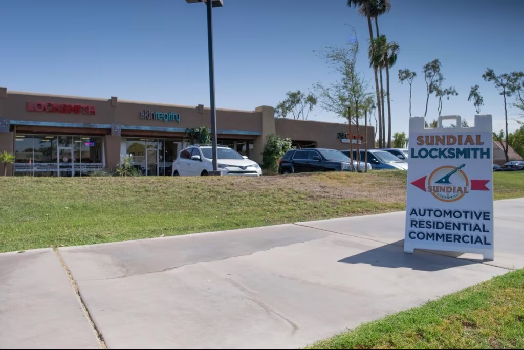 A Sundial Locksmith roadside sign advertising automotive, residential, and commercial services stands in front of a strip mall location on a sunny day.