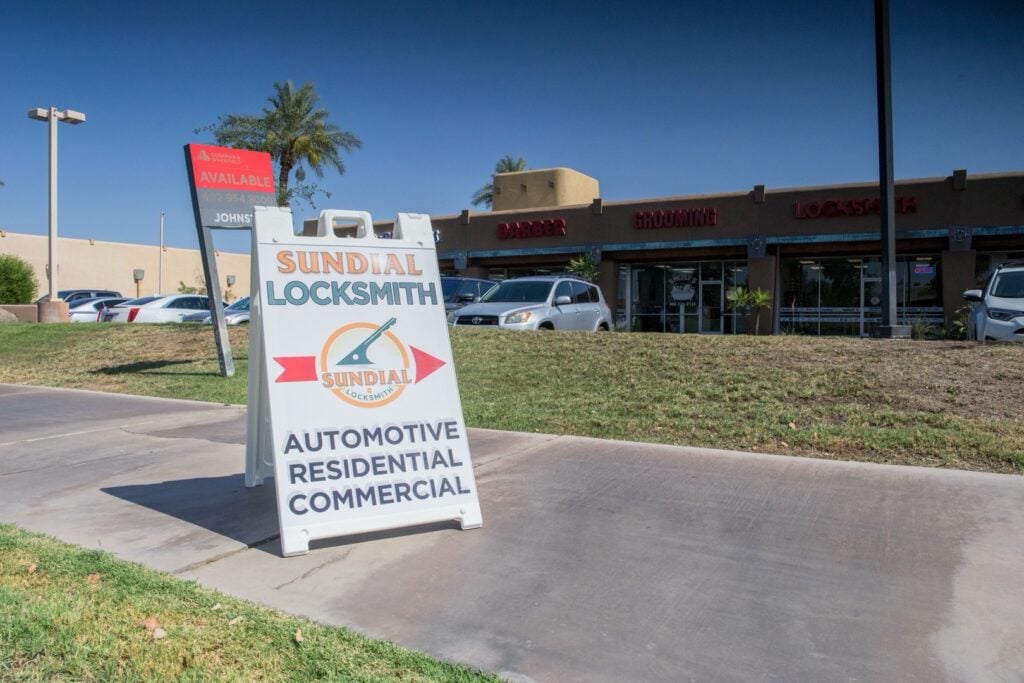 A-frame sidewalk sign for Sundial Locksmith advertising automotive, residential, and commercial services in front of a strip mall on a sunny day.