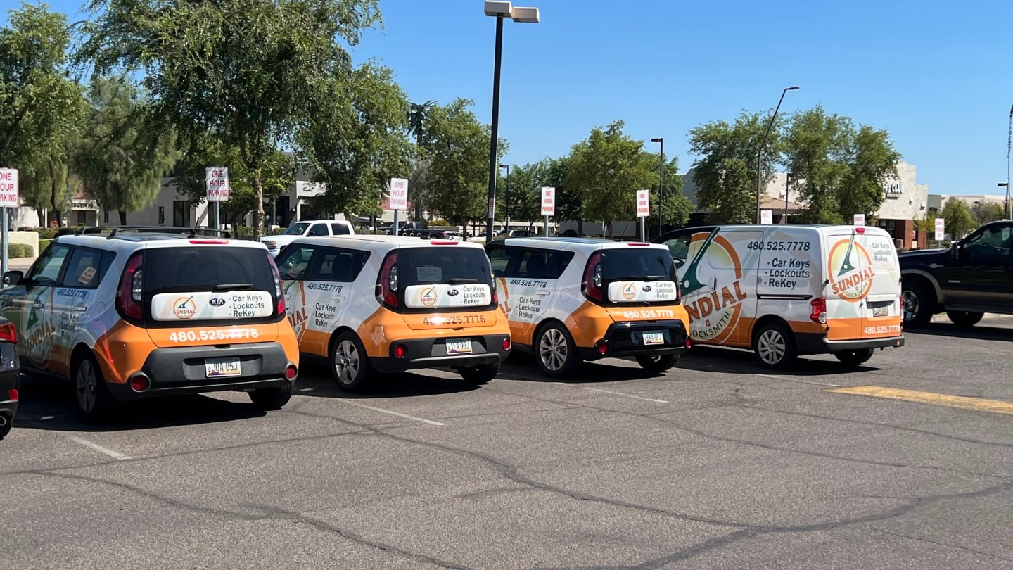 Four orange and white branded Kia Soul vehicles belonging to Sundial Locksmith parked in a row in a parking lot.
