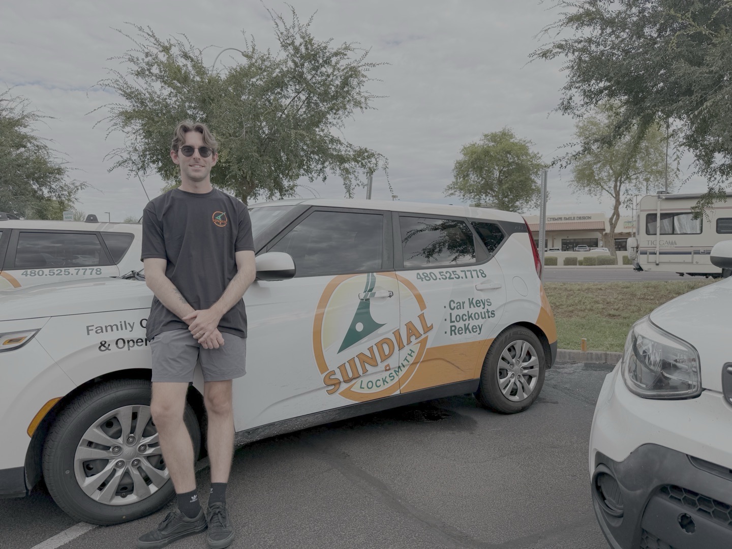 A young male Sundial Locksmith technician wearing sunglasses stands beside a white branded company car in an outdoor parking area.